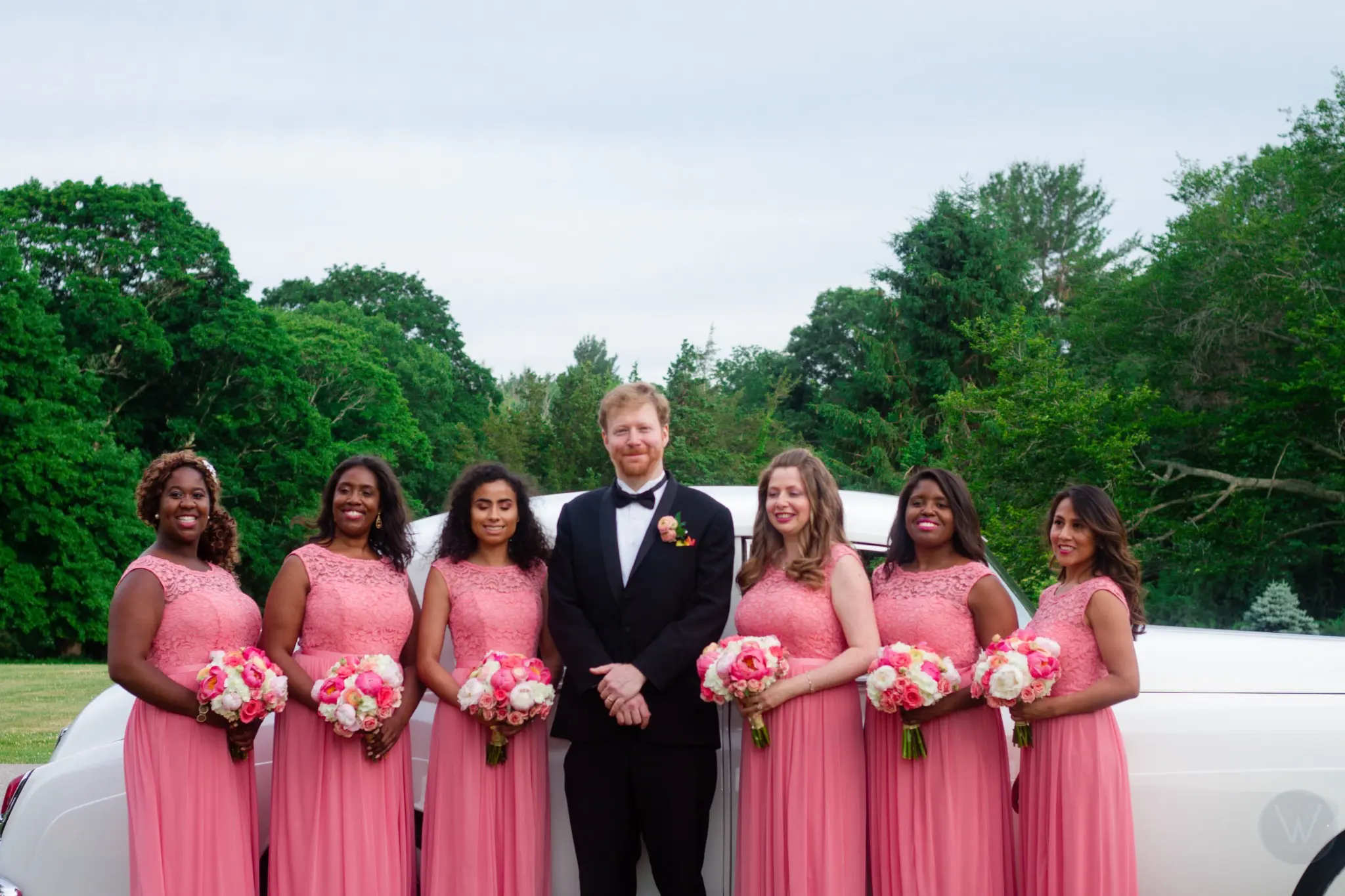 Bride, groom, and wedding party standing beside a white limousine